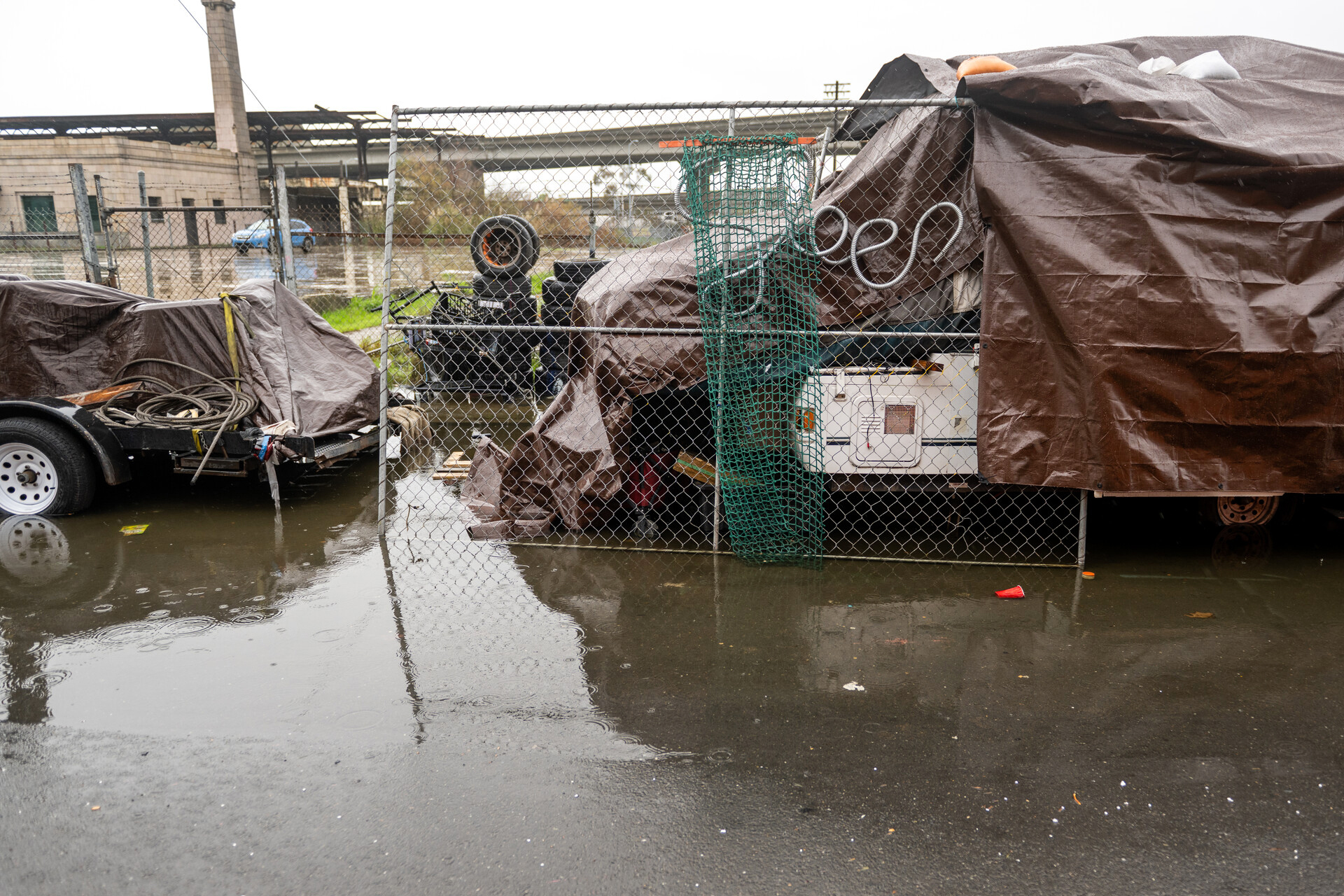 An outdoor shot of an unhoused person's encampment site. A white trailer is covered in brown tarps as pools of water start to form in front of the place. Piles of abandoned tires are in the background amid a gray sky.