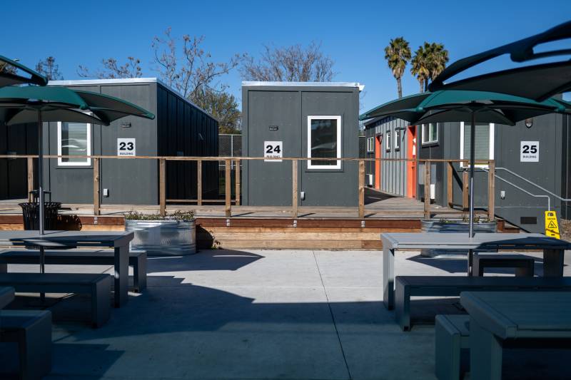 Neat rows of shipping containers have been converted into housing with white windows. An outdoor courtyard with gray picnic benches and sun umbrellas decorate the front.