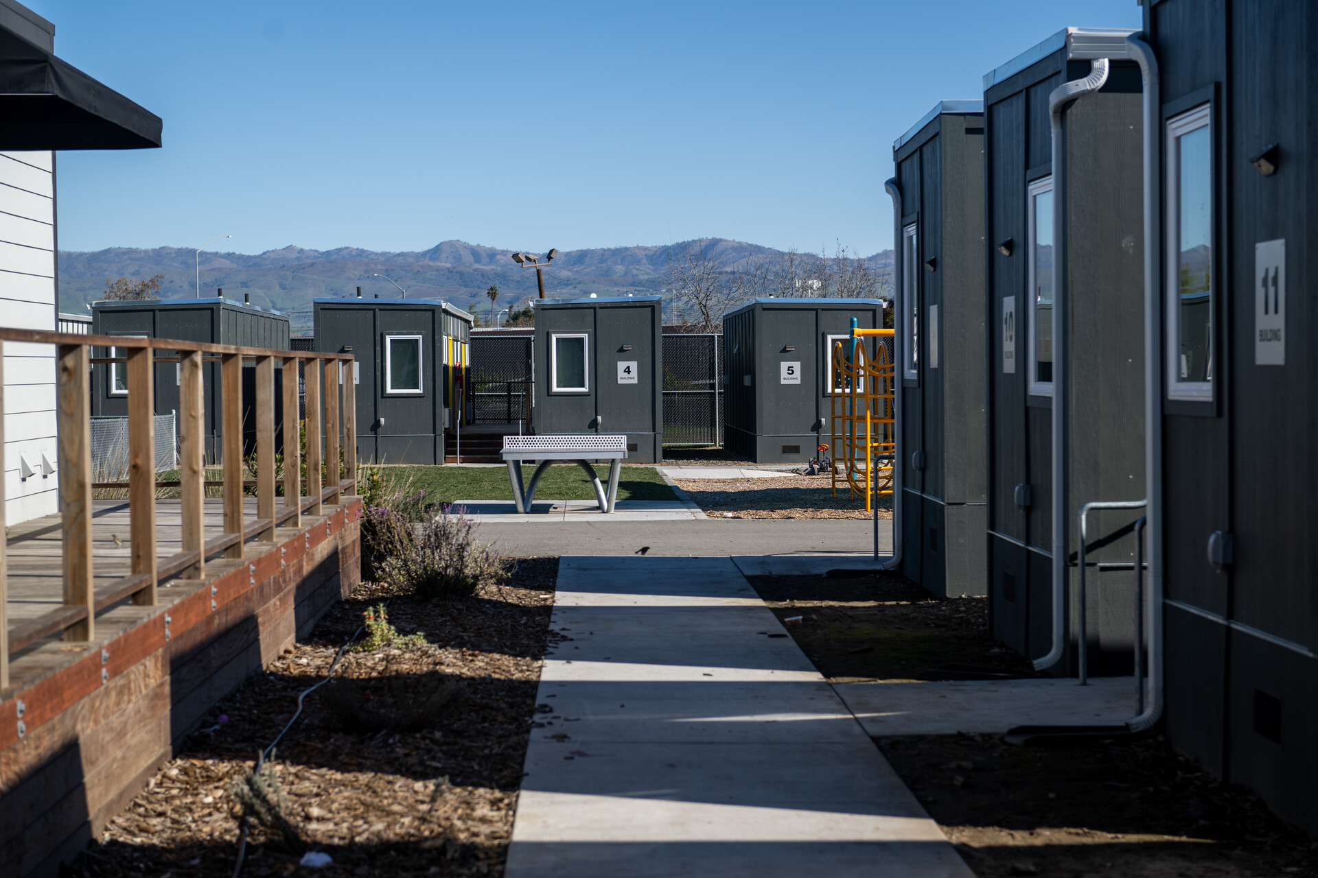 Neat rows of gray shipping containers with a singular white window in each line a sidewalk with well-manicured flower beds filled with brown bark. A mountain range is seen in the background.