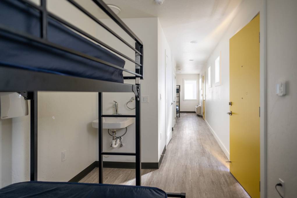 A bunk bed with black frame and mattresses covered in navy sheets is inside a small shipping container that was converted into temporary housing. A yellow door and a sink are seen just behind the bunk bed.