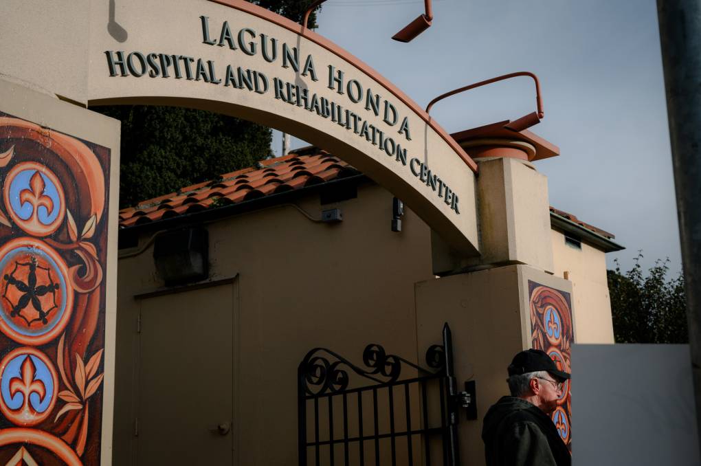 The outside of Laguna Honda nursing home has its namesake over an archway in green letters. Pillars on either side have red, orange and blue artwork painted on them. A man with gray hair wear a black baseball cap and a dark hooded sweater stands out front.