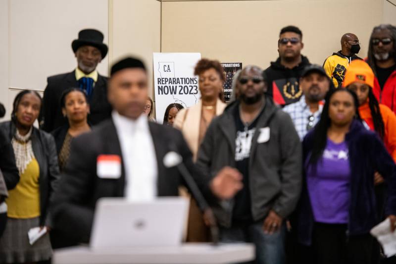 A group of Black men and women with white and black reparations sign behind them stand facing a man behind a table with a microphone and laptop.