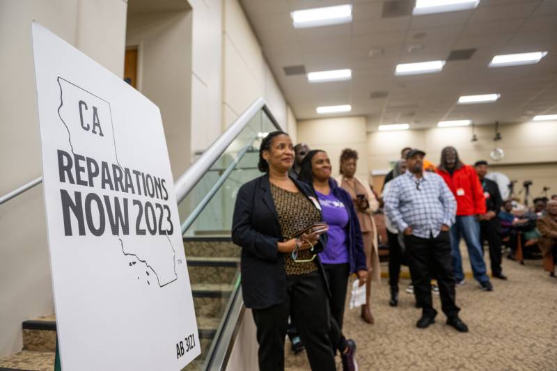 A black and white sign reads, "Reparations Now 2023" and a line of people are to the right of it in business-casual clothing.