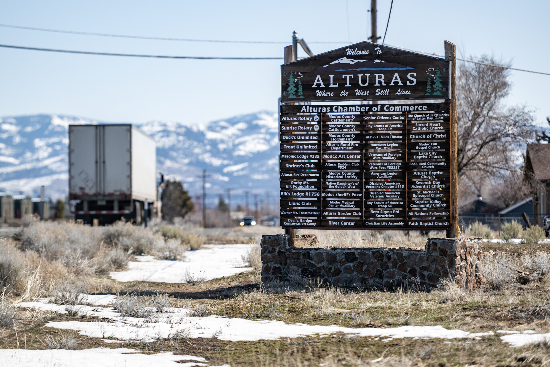 A large, wooden sign in the middle of yellow grass and brush reads "Welcome to Alturas: Where the West Still Lives." A snowy mountain range is seen in the background and a semi truck drives down a country road. Snow is melted on the ground and telephone poles dot the road.