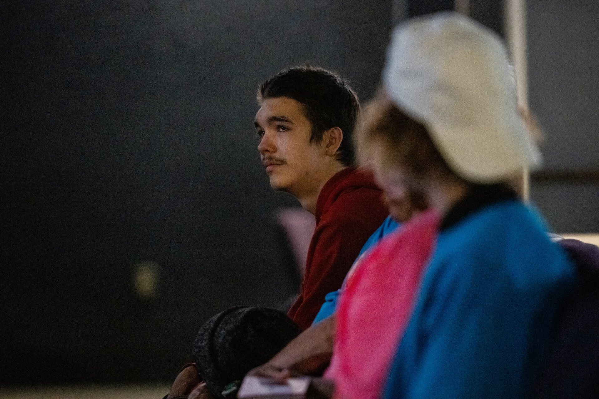 A young man with short, brown hair and a red hoodie sits in a row among family inside a church where a funeral is being held for his partner and his baby who were killed in a mass shooting. His face is heartbroken.