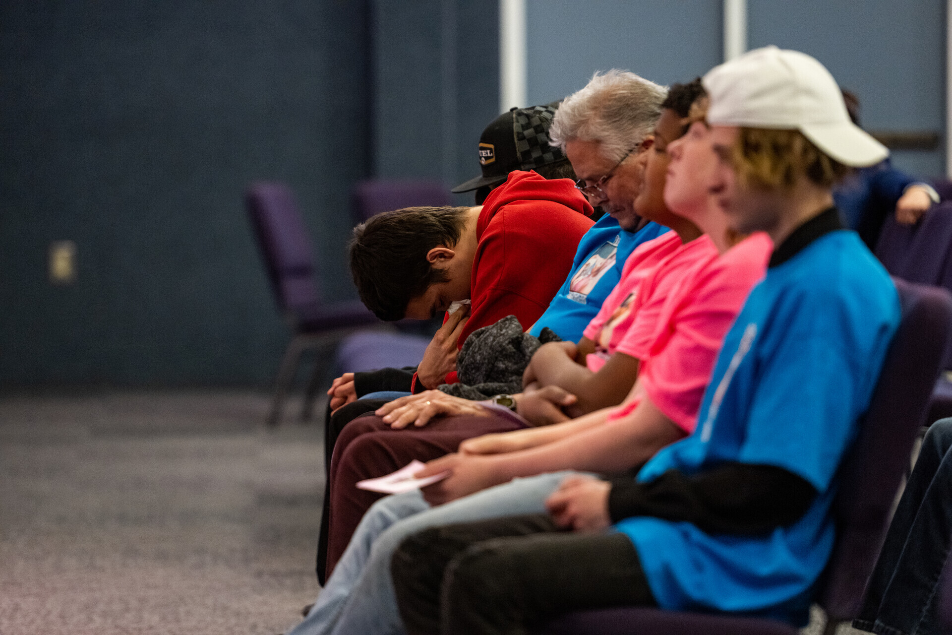A row of mourning family members, from teens to older males, sit inside a church. One bows their head with a sad expression as they listen to speakers during a funeral service for their loved ones who've died.