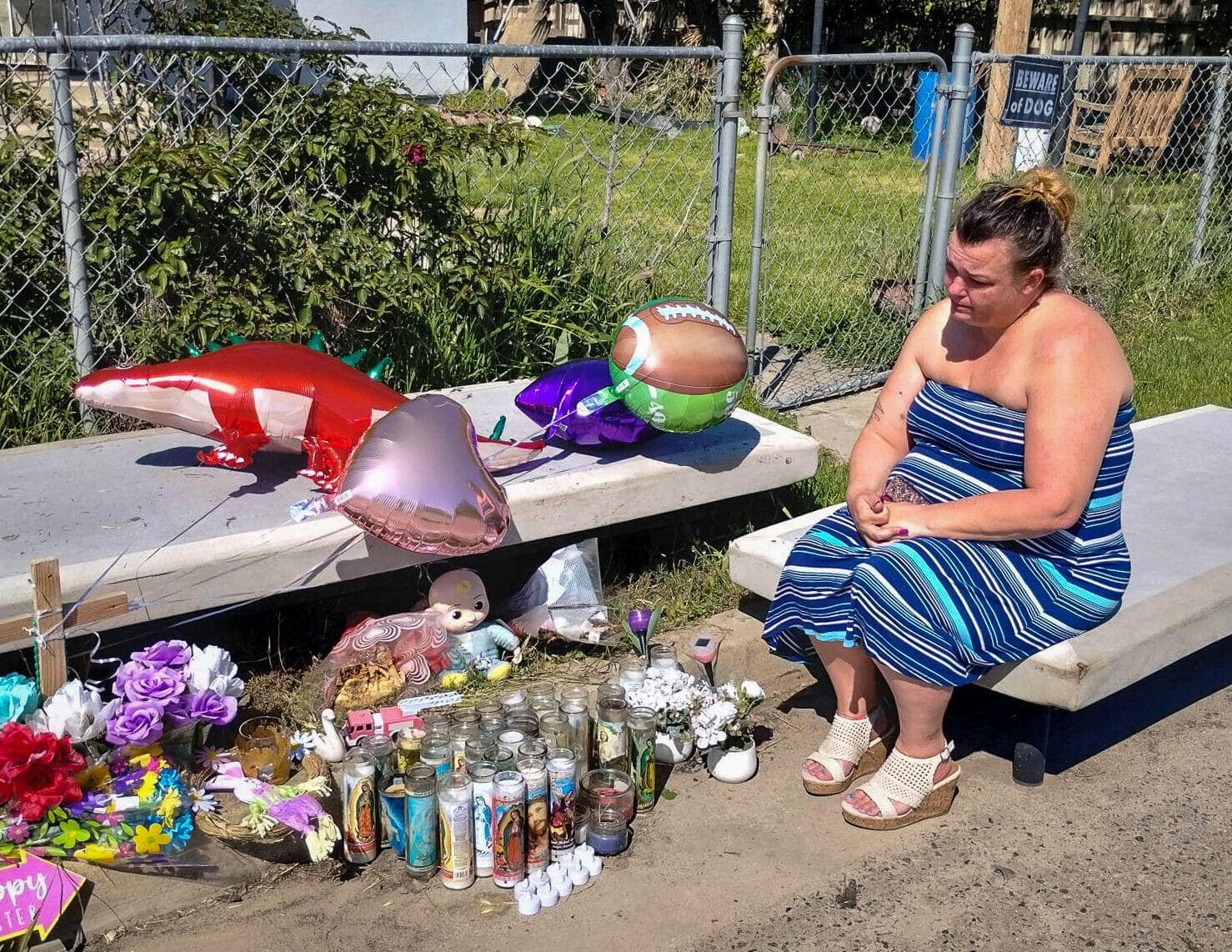 A woman sits at a memorial site where her loved ones were shot and killed. Votive candles, purple and red bouquets, balloons, and a wooden cross are all positioned on a dirt sidewalk in front of a chain link fence. The woman sits on an abandoned mattress as she stares solemnly at the display.