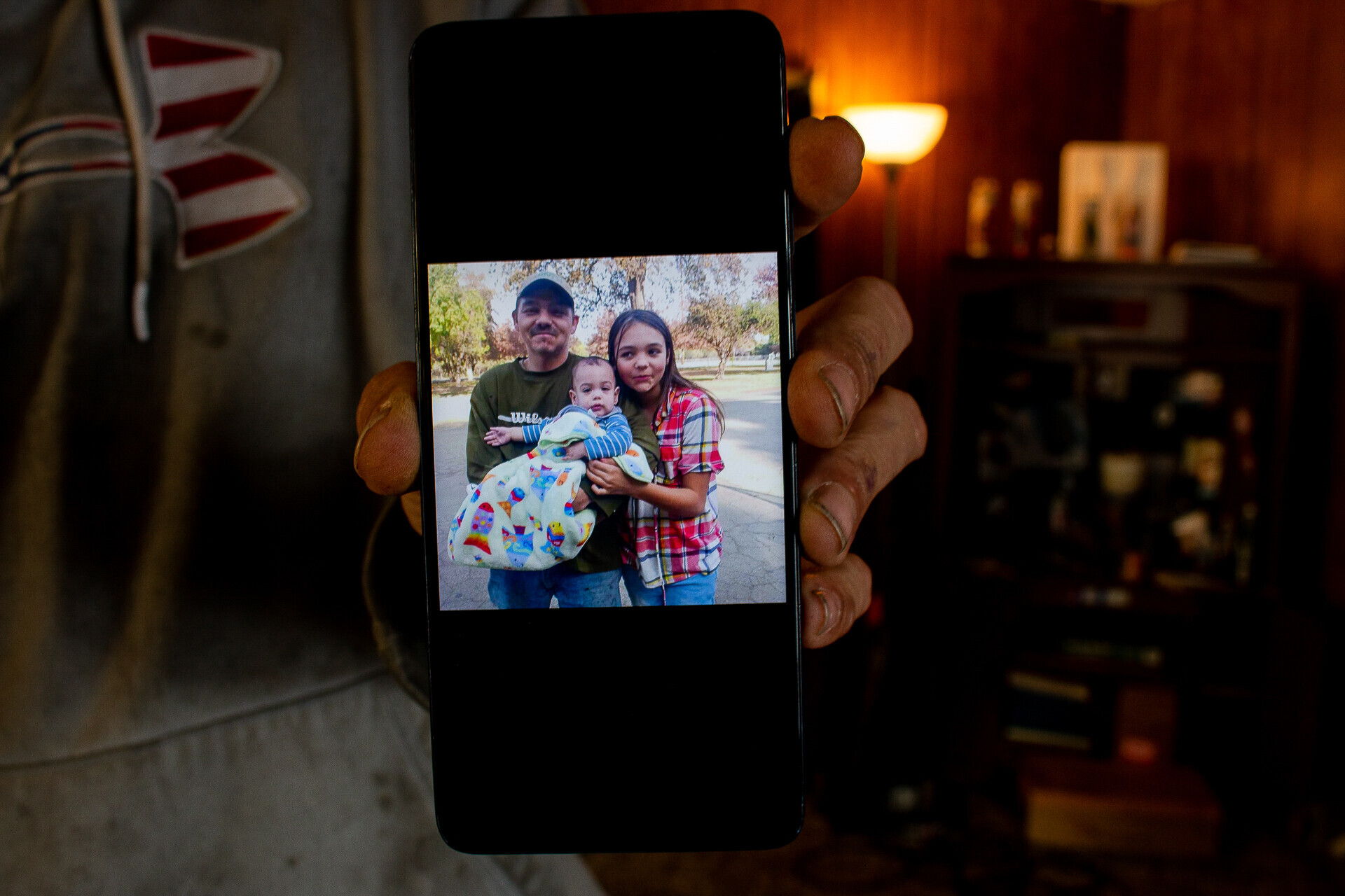 A man's hand holds a cell phone that displays a photo of himself with a teenage girl and her baby boy who clutches a baby blanket.