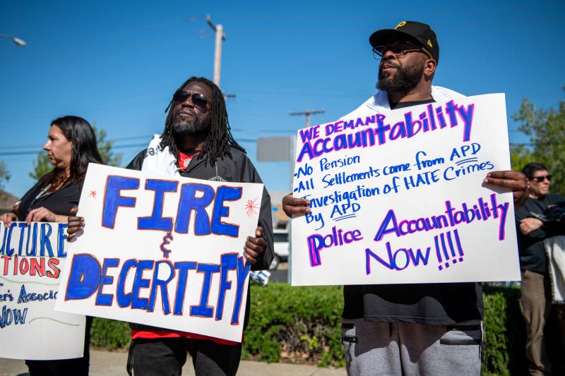 Two Black men and one woman hold signs at a protest calling for police accountability.