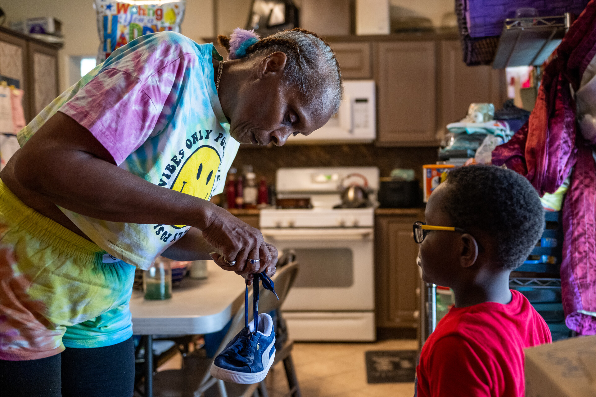 An at-home child care worker in a rainbow T-shirt helps a little boy in a red T-shirt untie a knot in his black sneaker laces.