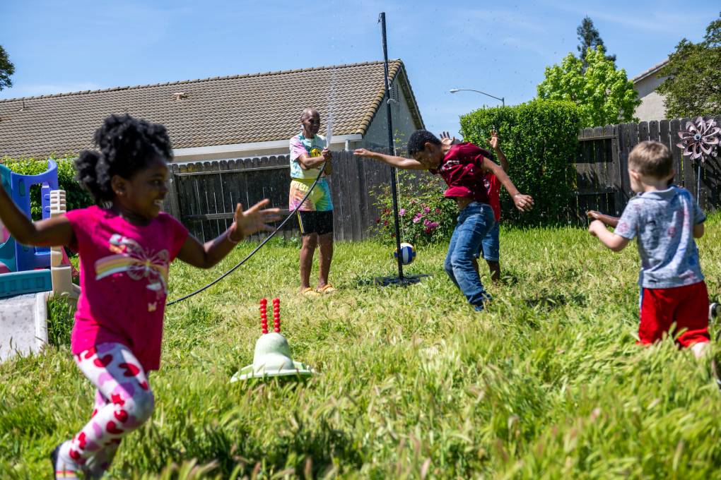 Children run around a tether ball pole amid long blade of green grass. Daycare aides are seen smiling nd playing along.