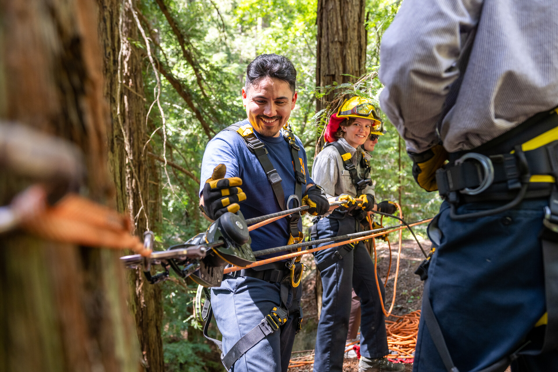 A Latino trainee smiles as he works the rope controls with a female trainee looking on and smiling in a forest setting.