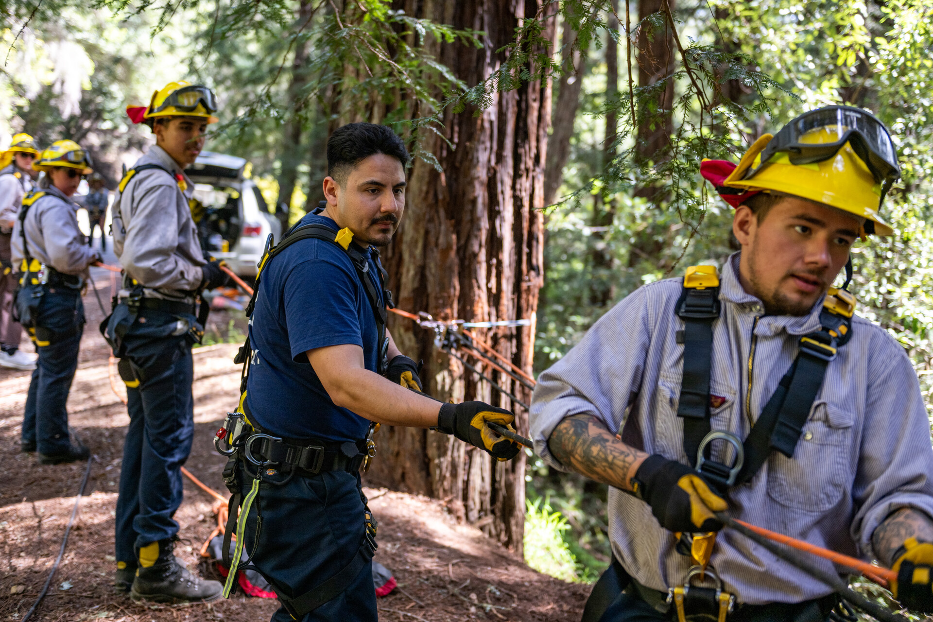 A group of trainees wearing firefighting gear hold a rope as they stand in line behind each other, all but one wearing yellow helmets and goggles, trainees are male and female of different ethnicities.