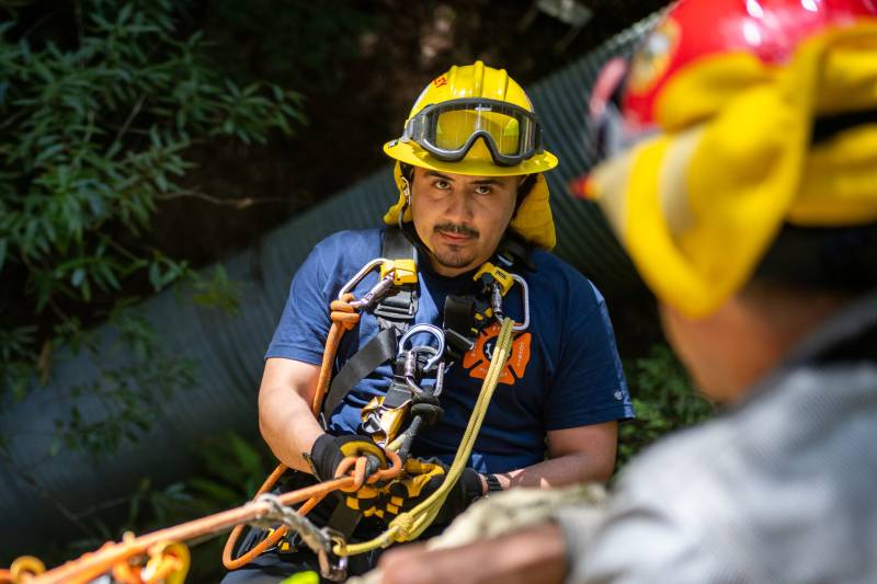 A Latino man wearing fire gear and helmet with goggles looks up at a fire instructor while rappelling down a cliff.