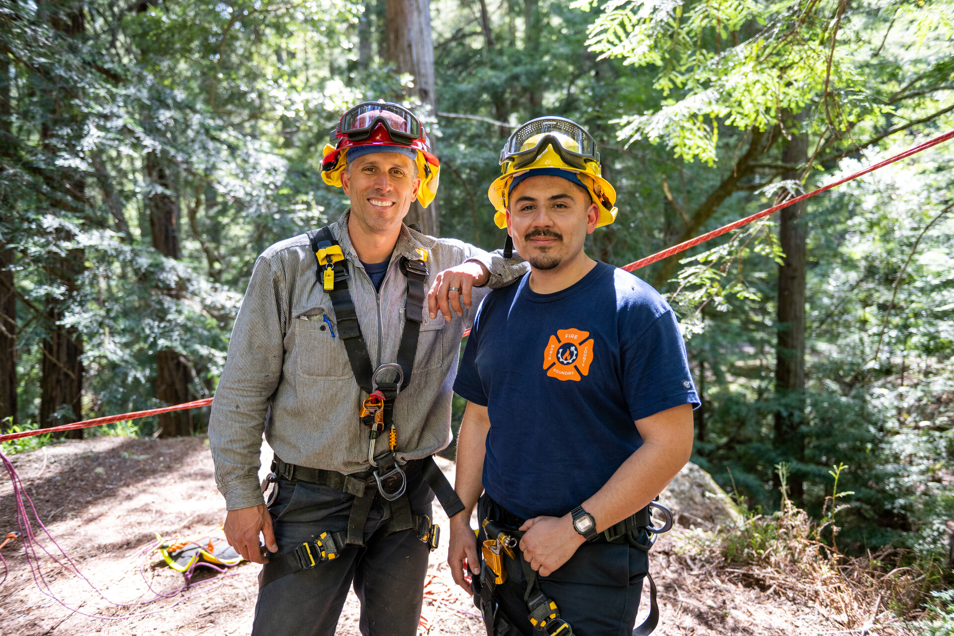 A white man and a Latino man wearing fire gear and helmets with goggles smile at the camera in the woods with a rope running behind them.