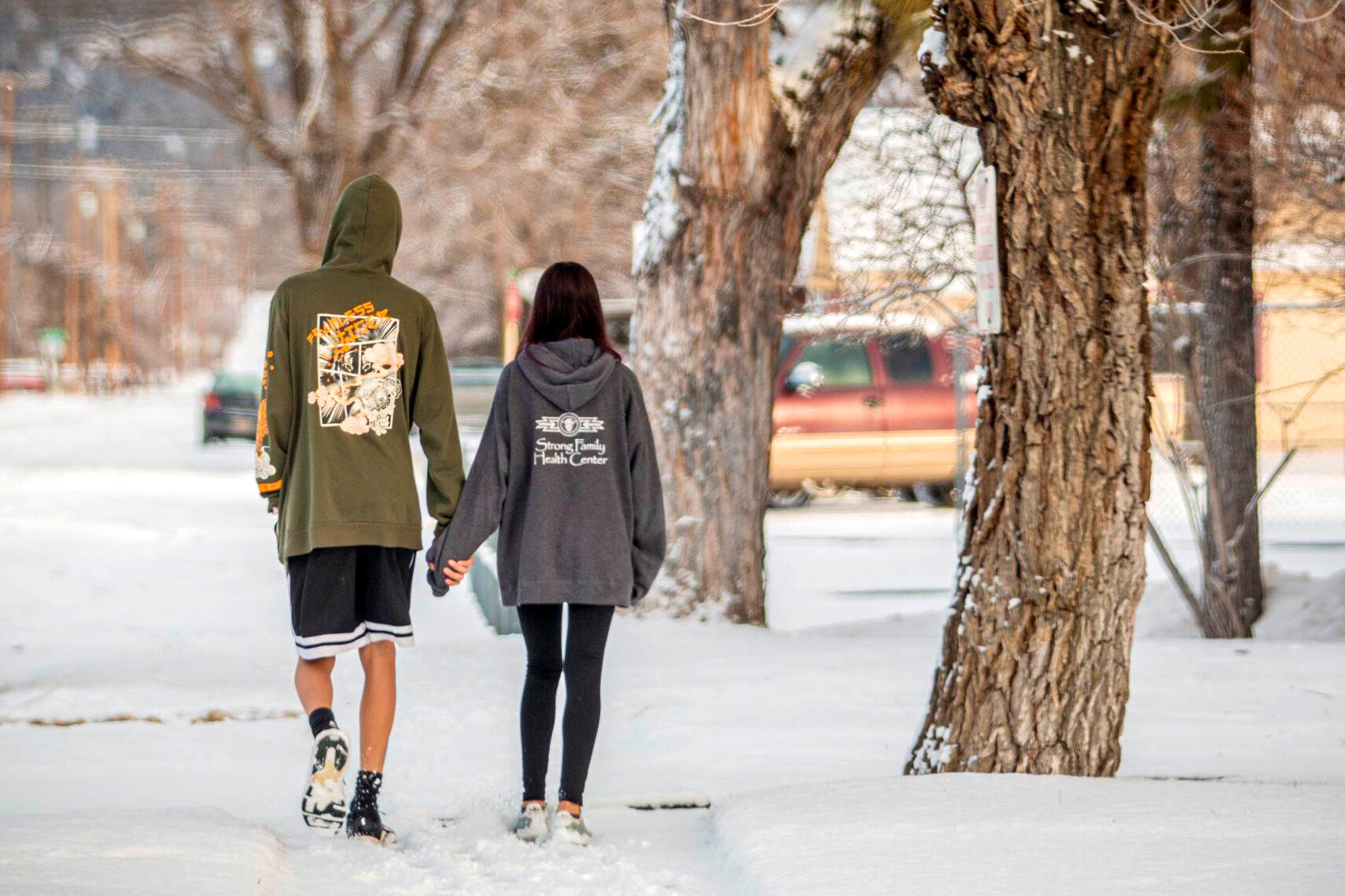 A young couple walks in the snow holding hands with their backs toward the camera.