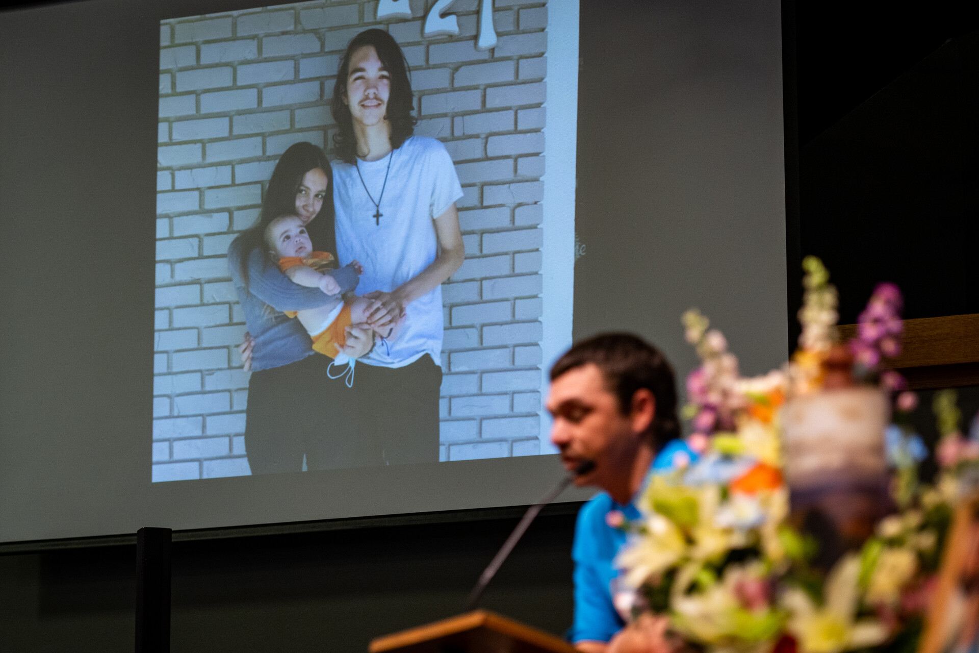 A man at a podium inside a church memorial service speaks with a projected photo of a young couple and their baby behind him.