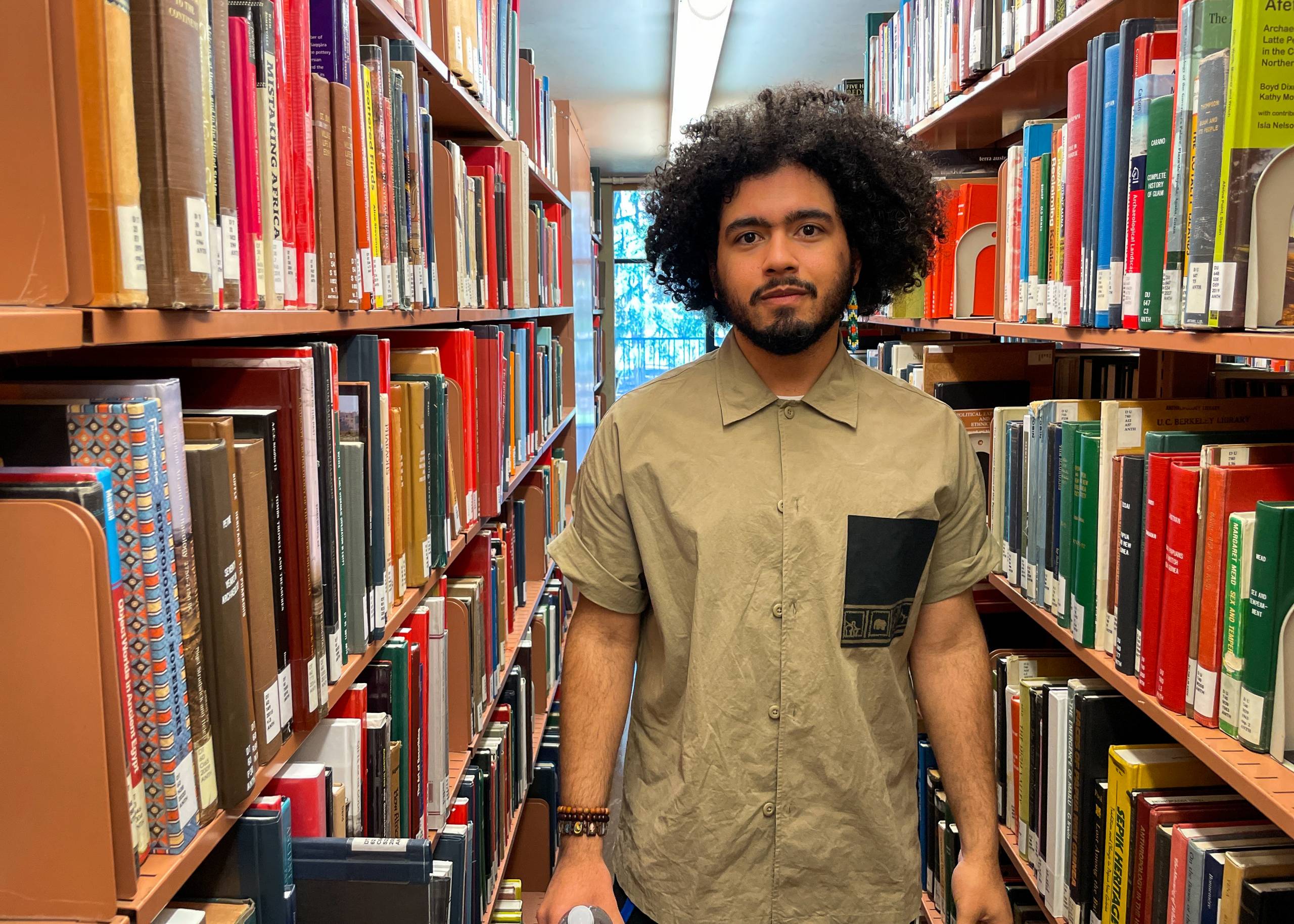 A young man with bushy hair poses among the stacks of a library. 