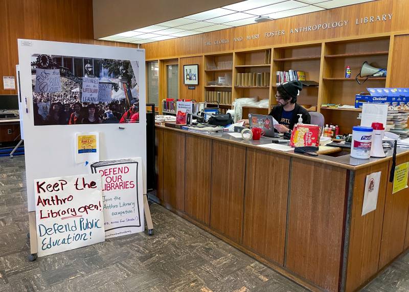 A man sits behind a library circulation desk. In front of him is a large sign board protesting the library's closure.