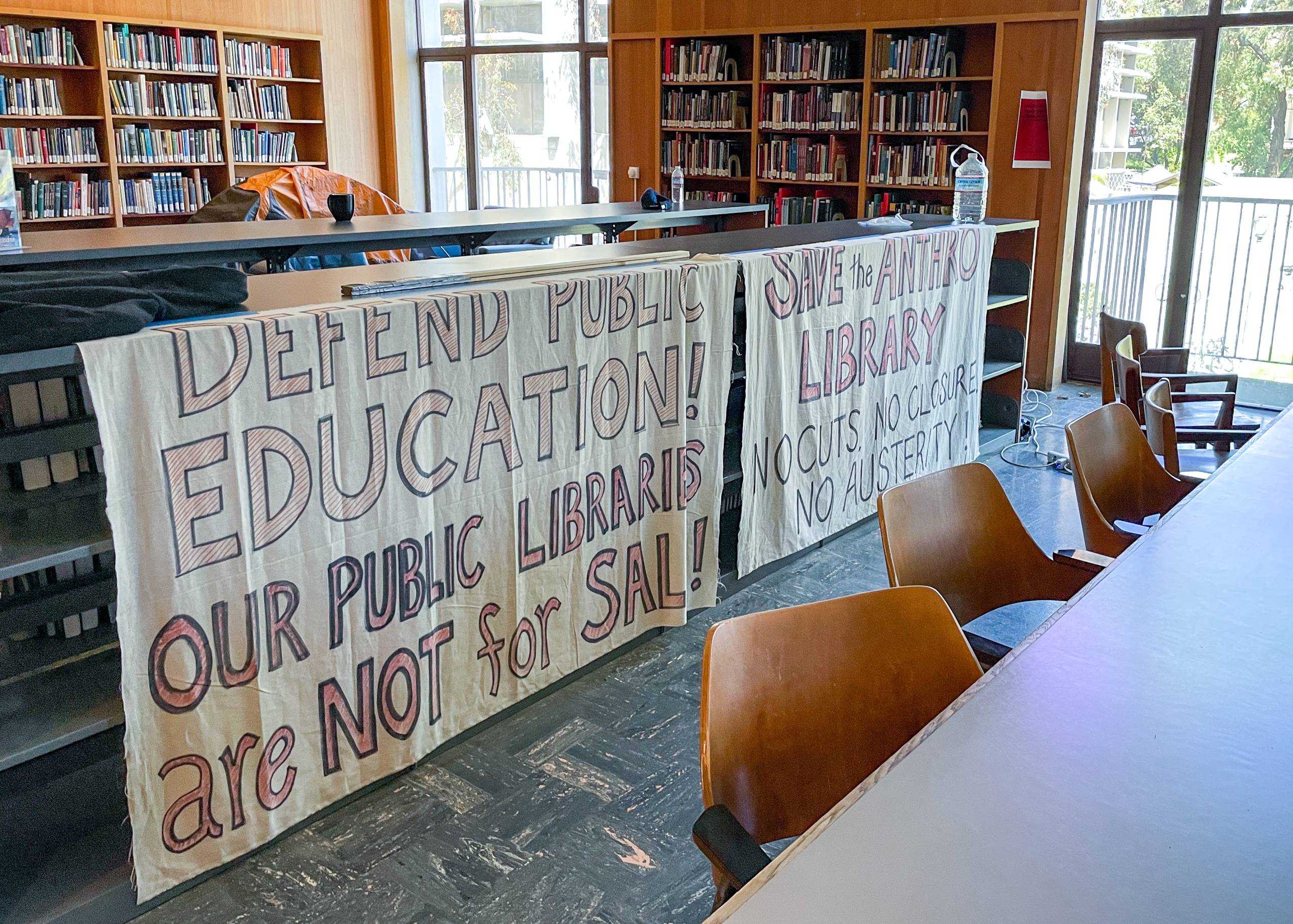 Cloth protest banners hanging from low shelves in the library, that say, 'Defend Public Education' and 'Save the Anthropology Library.'