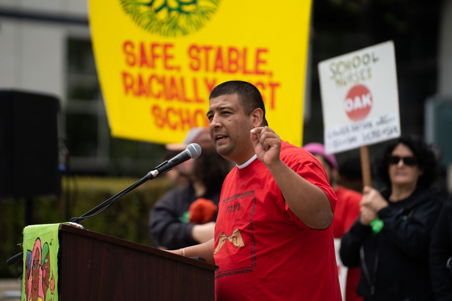 A man in a red T-shirt with dark hair speaks from a podium as large signs behind him read, "Safe, Stable, Racially Just Schools."