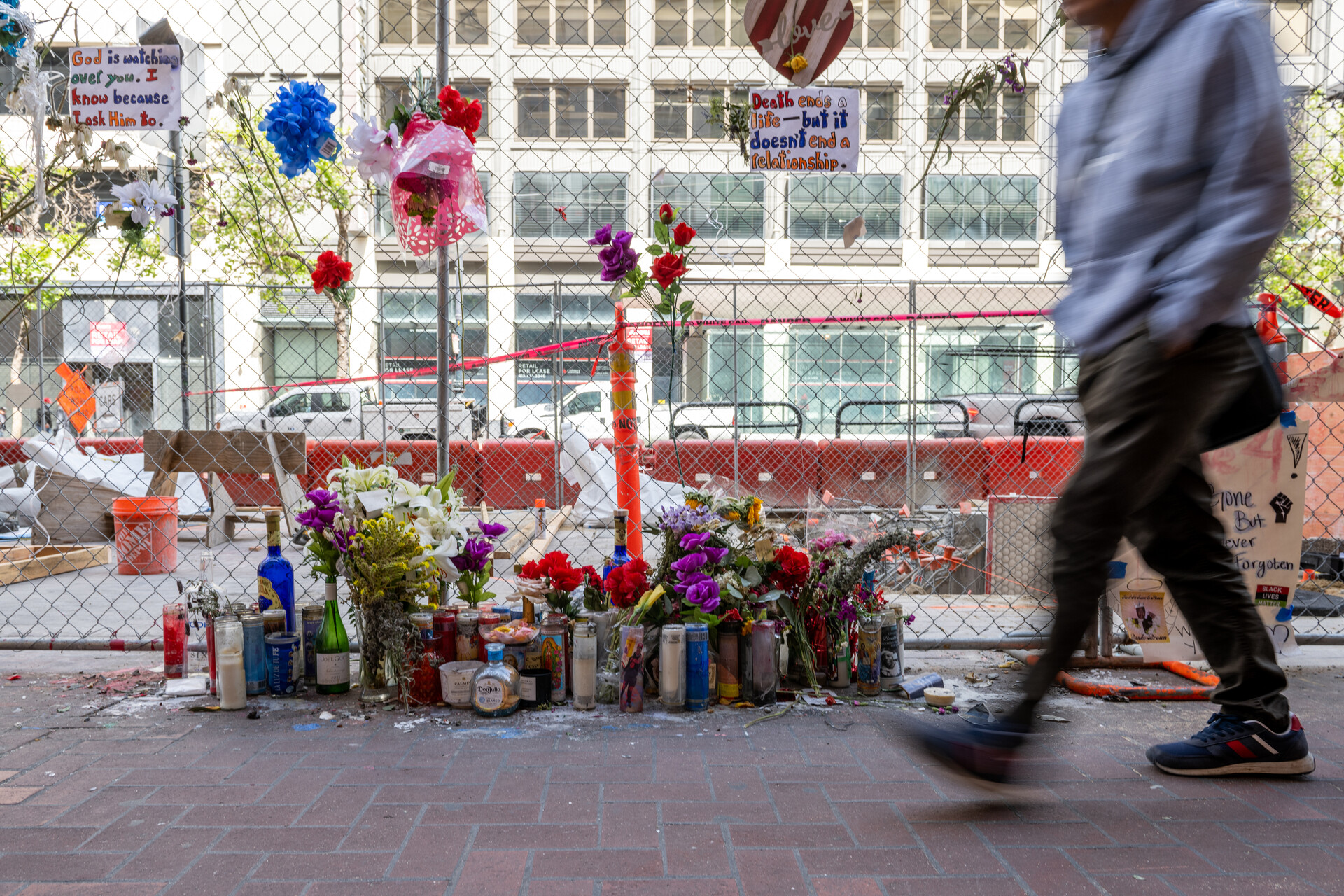 candles, photos and posters sit against a chain link fence in the background as the blurry feet of a pedestrian walk past in the foreground