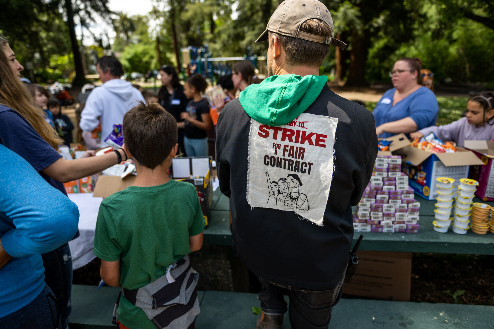 A little boy in a green T-shirt and a striped sweater wrapped around his wait stands next to a man with a black hoodie on with a back patch that reads, "Strike for a fair contract." Both of their backs face the camera. The two are standing at a picnic bench with neat stacks of juice boxes and snacks for kids.