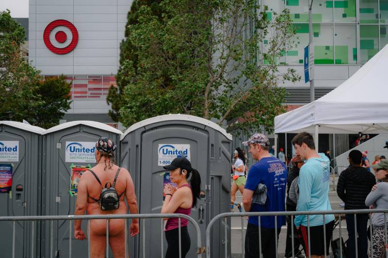 Several people in athletic clothing wait in line to use portable toilets. One man waits completely naked, except for a small backpack.