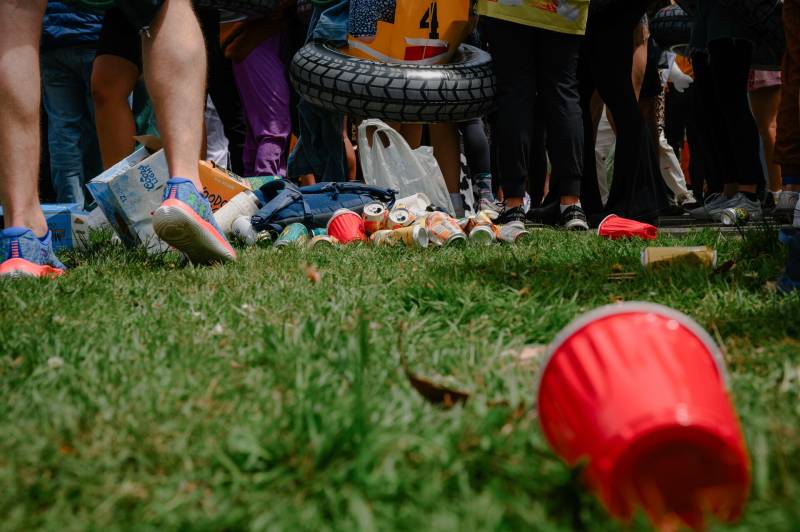 Low angle image taken from the grass. A red plastic cup lays in the foreground, while a piles of cans and beer boxes lay at the feet of a group of people further away.