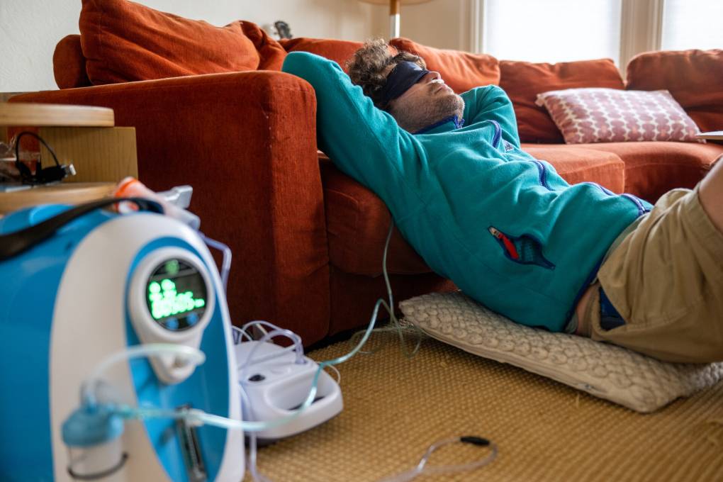 A white man wearing a turquoise sweater lies on a living room floor with his back propped against an couch with arms folded behind head and sleep mask on with a cord running from his nose to a portable oxygen machine in the foreground.