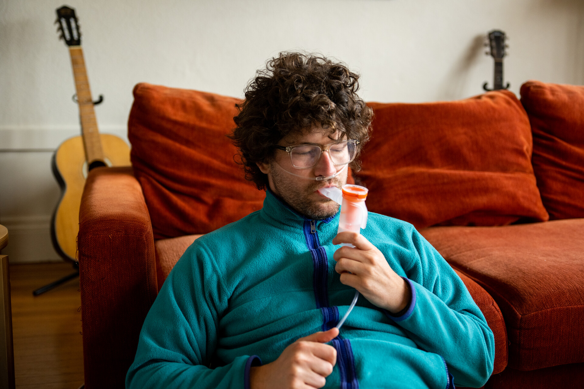 A white man with light brown curly hair and eyeglasses has a steam inhaler in his mouth as he laws reclines against a sofa in his living room.