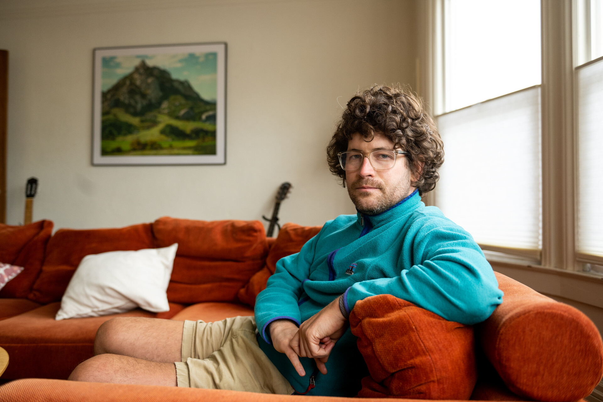 A white man with curly light brown hair and eye glasses sits on a couch with feet up and looks at the camera with head turned to the side.
