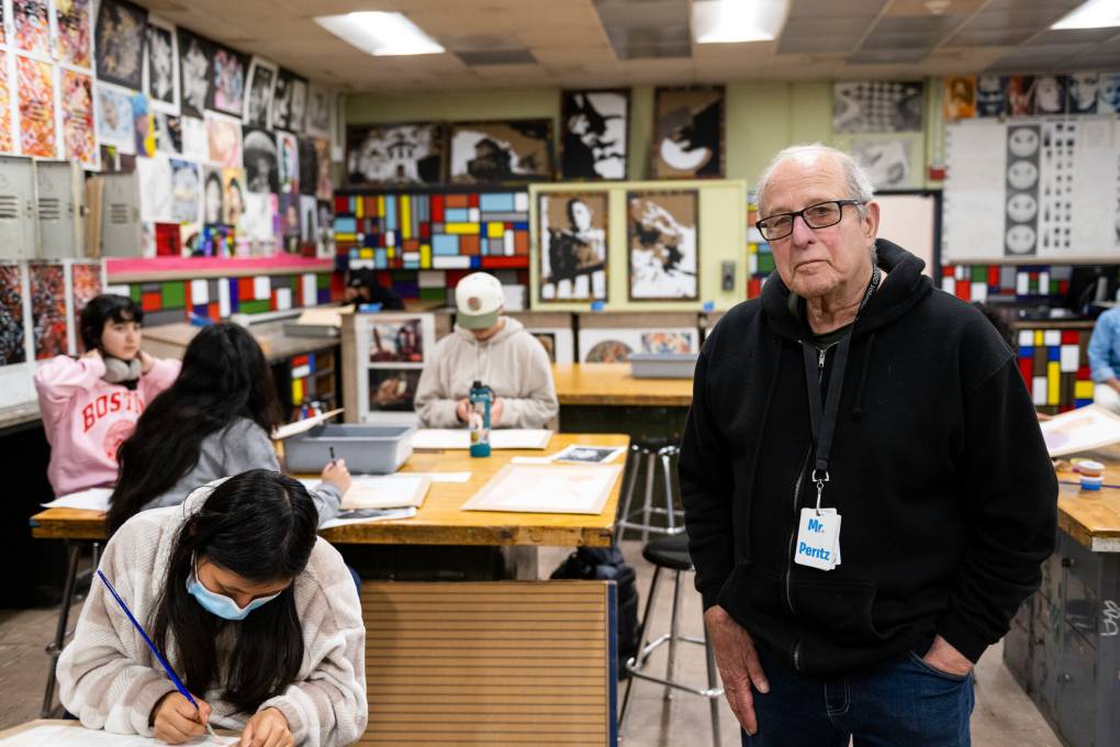 A white man with glasses in a black hoodie stands in a classroom with students seated at desk writing on pieces of paper.