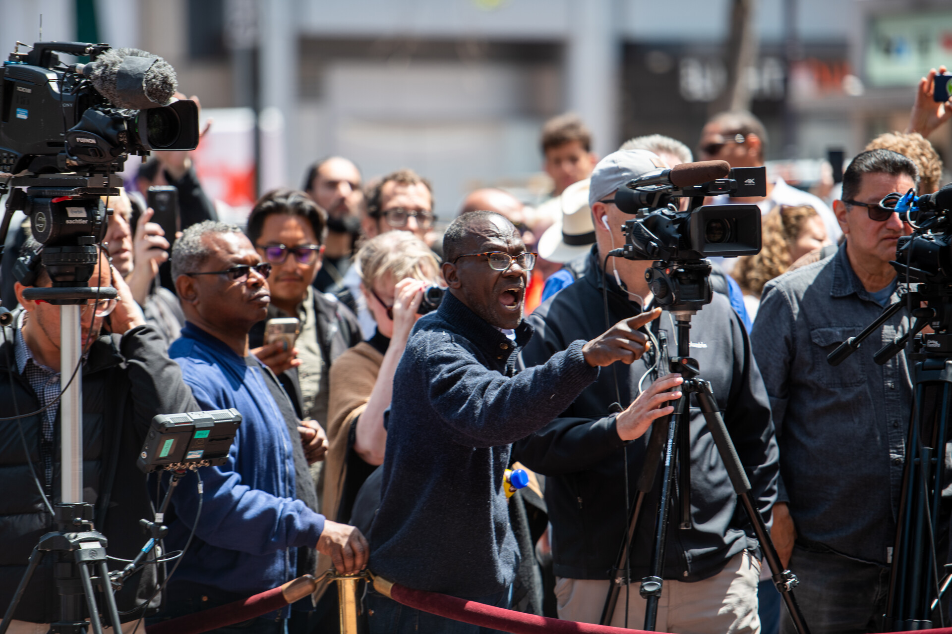 A middle-aged Black man stands behind a camera, in a crowd, shouting.