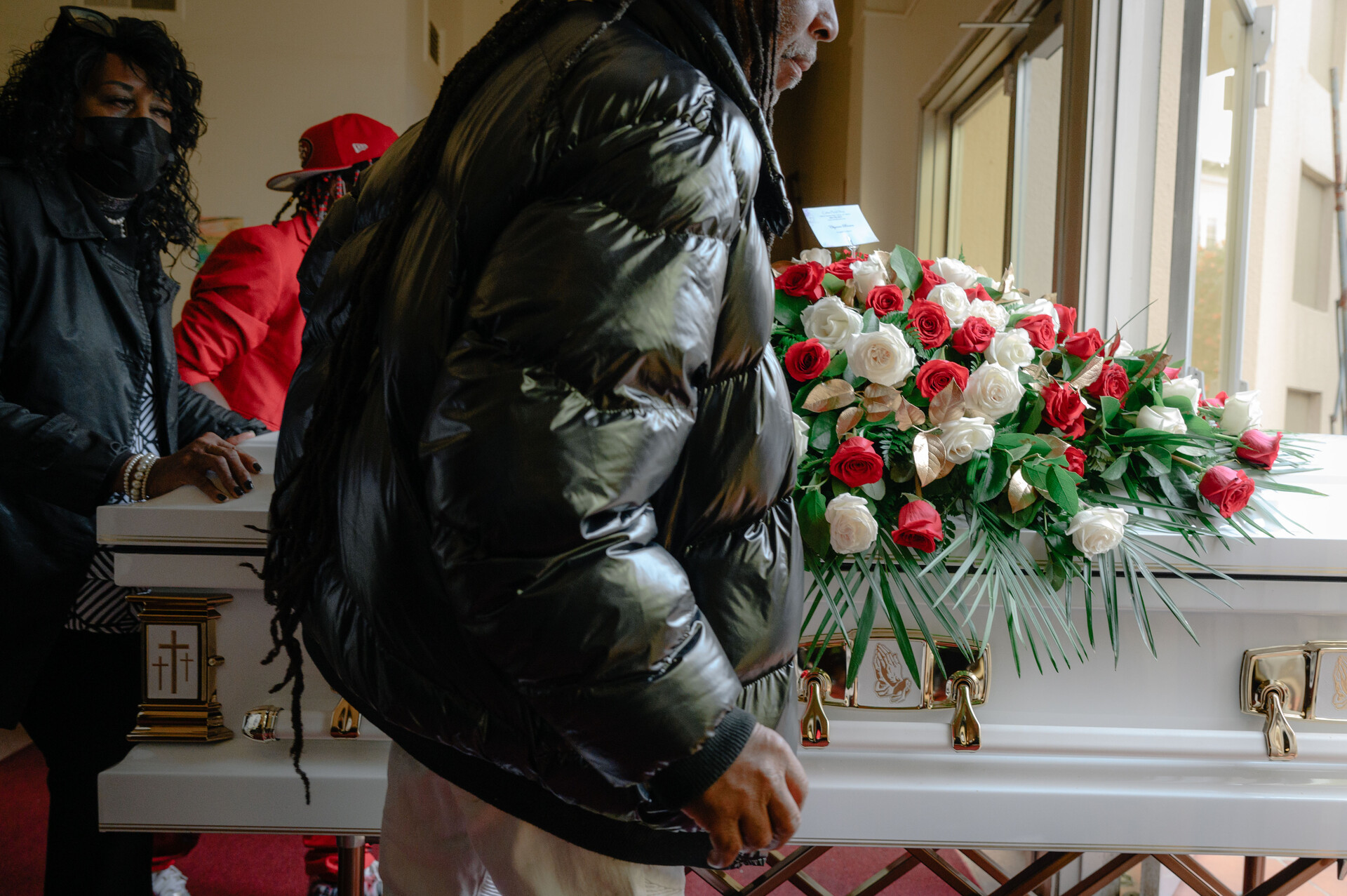 a man with dreadlocks, in a black coat, carries a white coffin covered in flowers, with two other men in the background also holding the coffin 