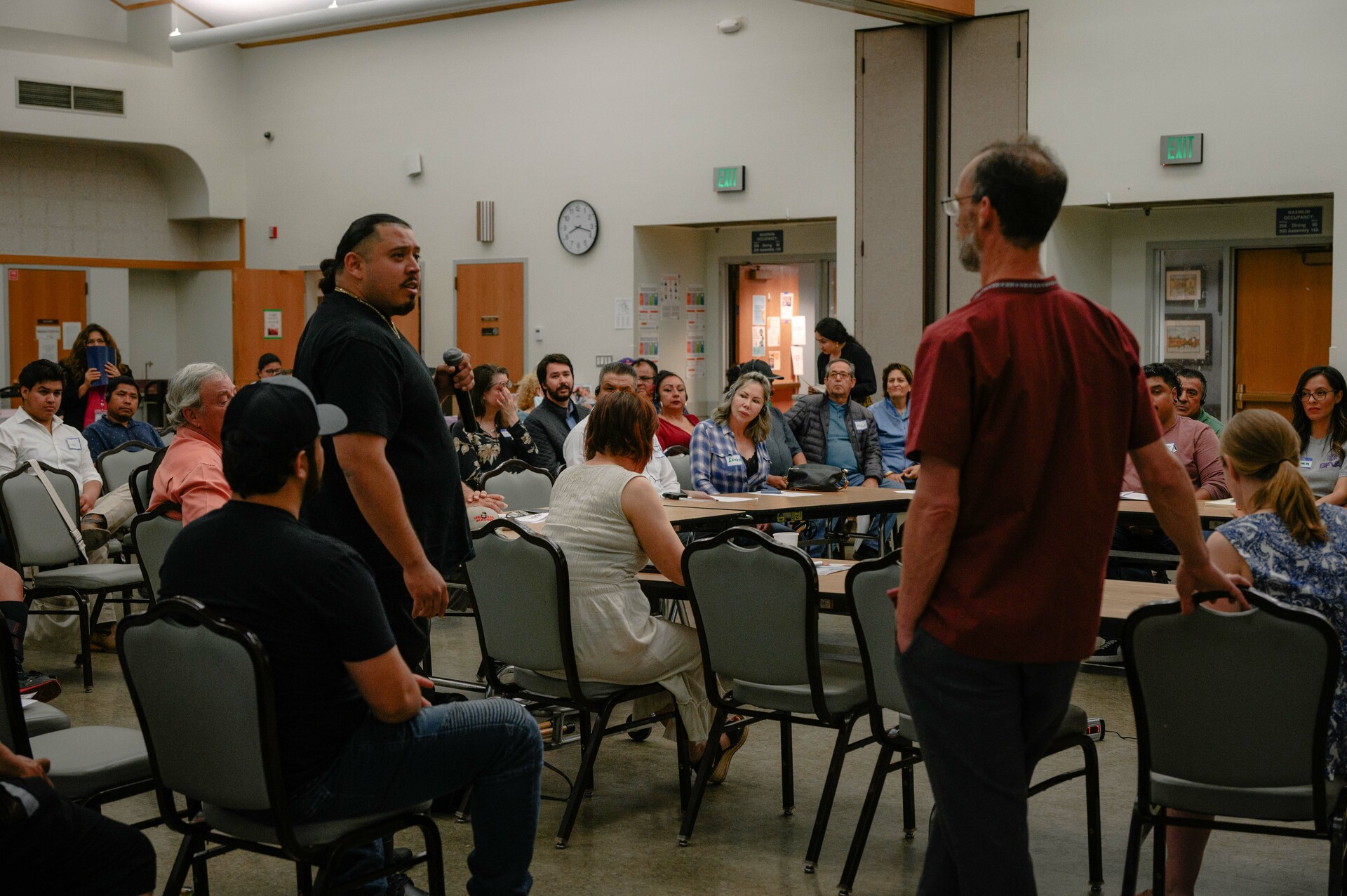 Two men are standing about six feet apart from each other in a crowded room with a seated audience. They attend a city meeting and are speaking to each other.