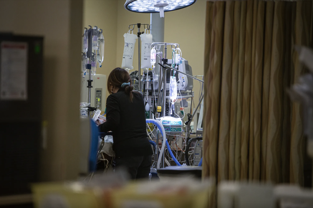 A woman wearing dark clothing with her back turned leans over someone in a hospital bed surrounded by medical equipment.