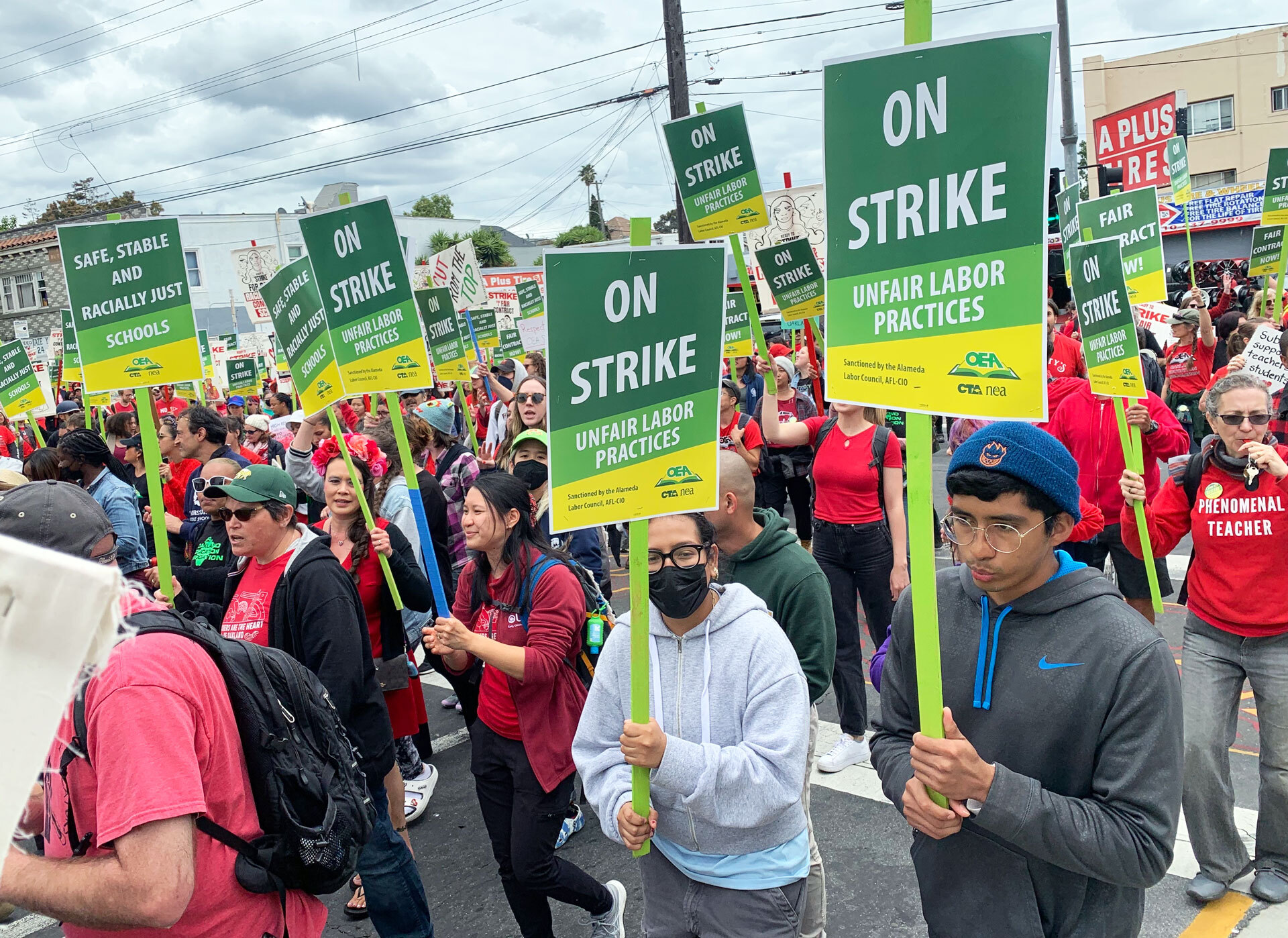 A huge crowd of protestors walk down a street in Oakland carrying green and yellow picket signs that read "On Strike Unfair Labor Practices." Many participants wear red and one woman has a hooded sweatshirt that reads "phenomenal teacher." Another sign reads "Safe, Stable and Racially Just Schools"