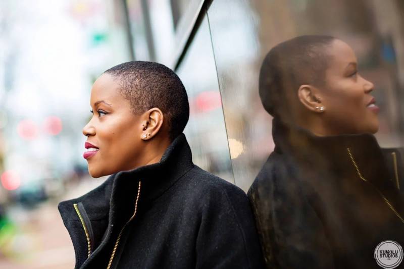 a Black woman with closely shaved hair, wearing a black coat, looks out of frame