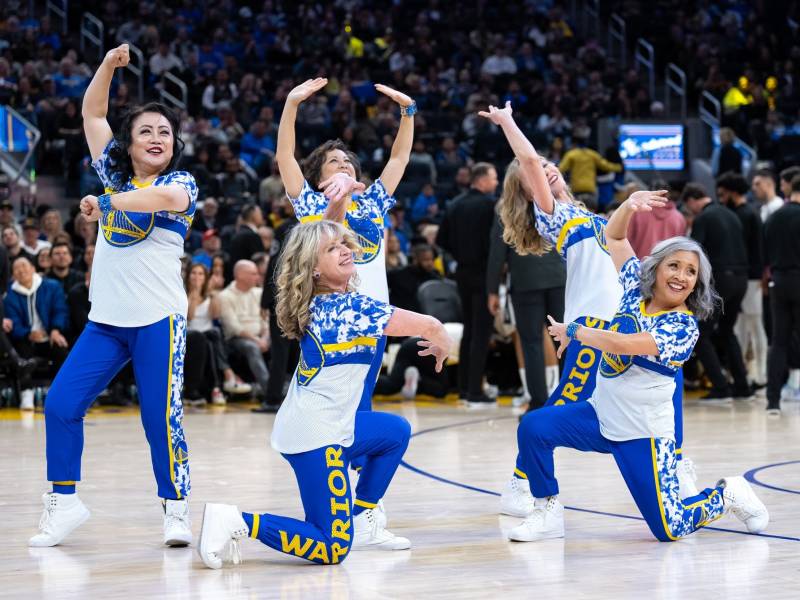 Five women wearing white, blue and yellow clothing pose in various positions in the middle of a basketball court with thousands of people sitting in the background.