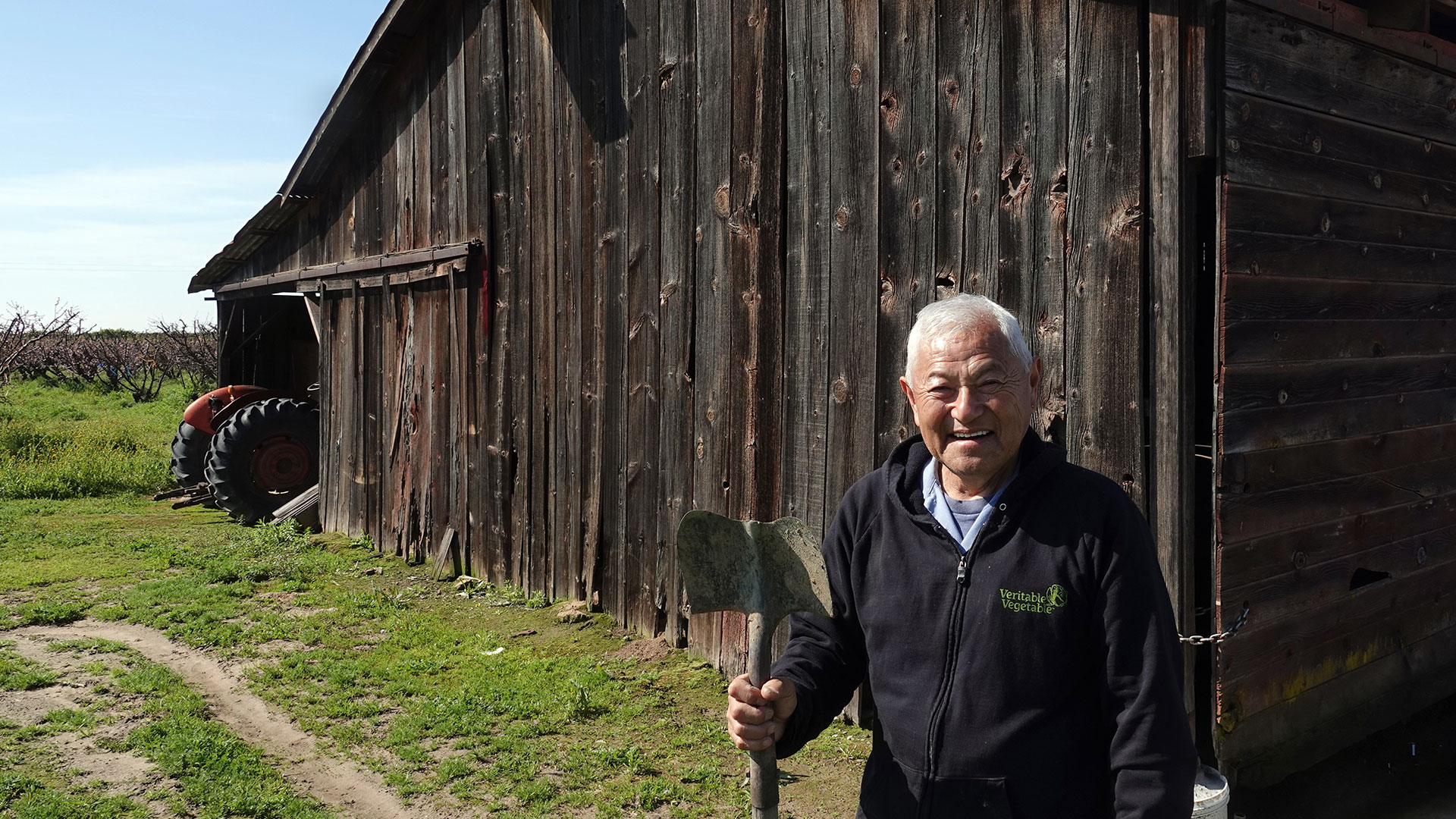 A Japanese American man with white hair and a black zip-up sweatshirt stands in front of a weathered wood barn holding a shovel. He is smiling broadly.