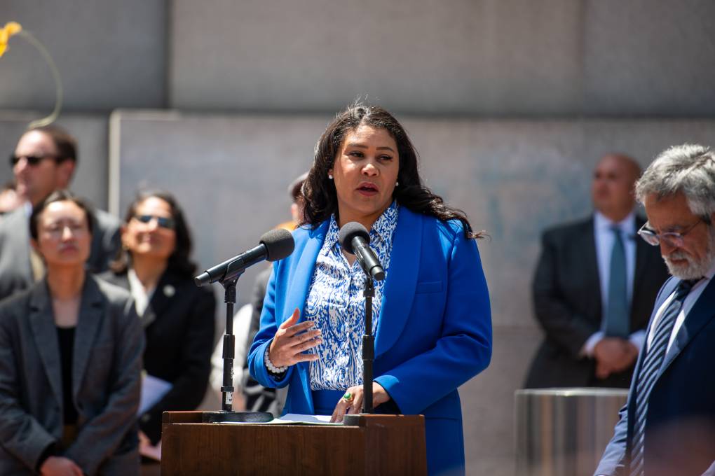 Mayor London Breed speaks outside at a lectern, next to a bearded white man
