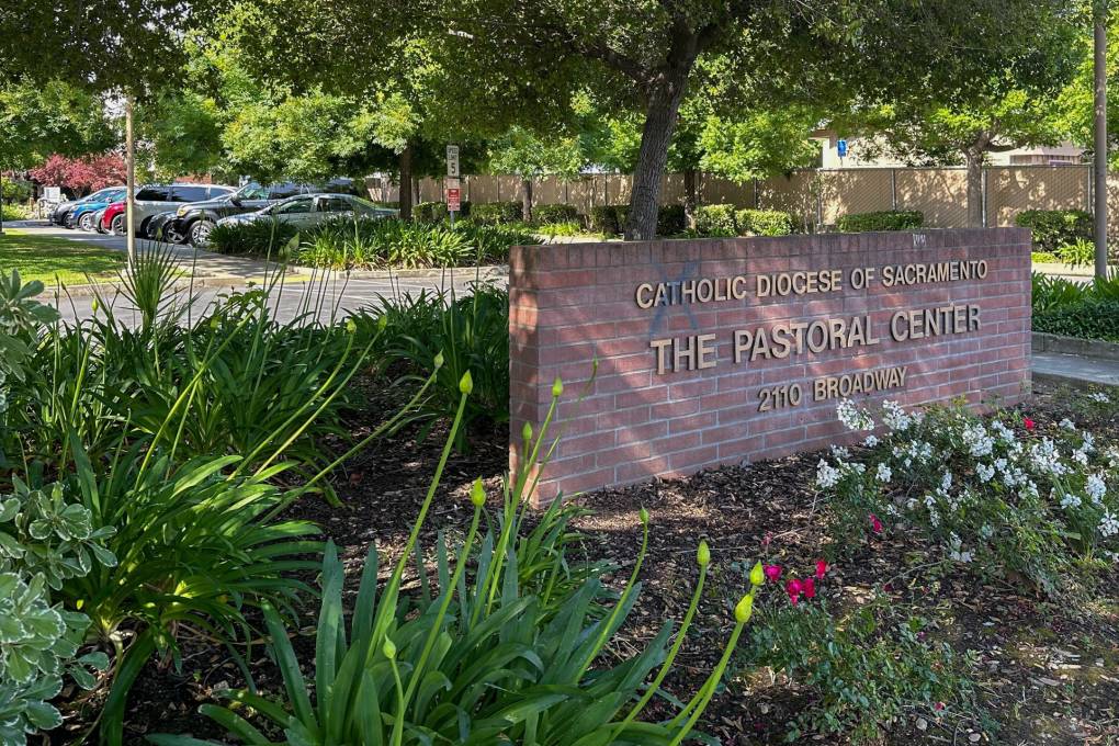 A brick, rectangular sign with gold letters surrounded by brown bark, white and red flowers and green bushes reads, "Catholic Diocese of Sacramento: The Pastoral Center 2110 Broadway." It's daytime and a parking lot full of vehicles is seen in the background.