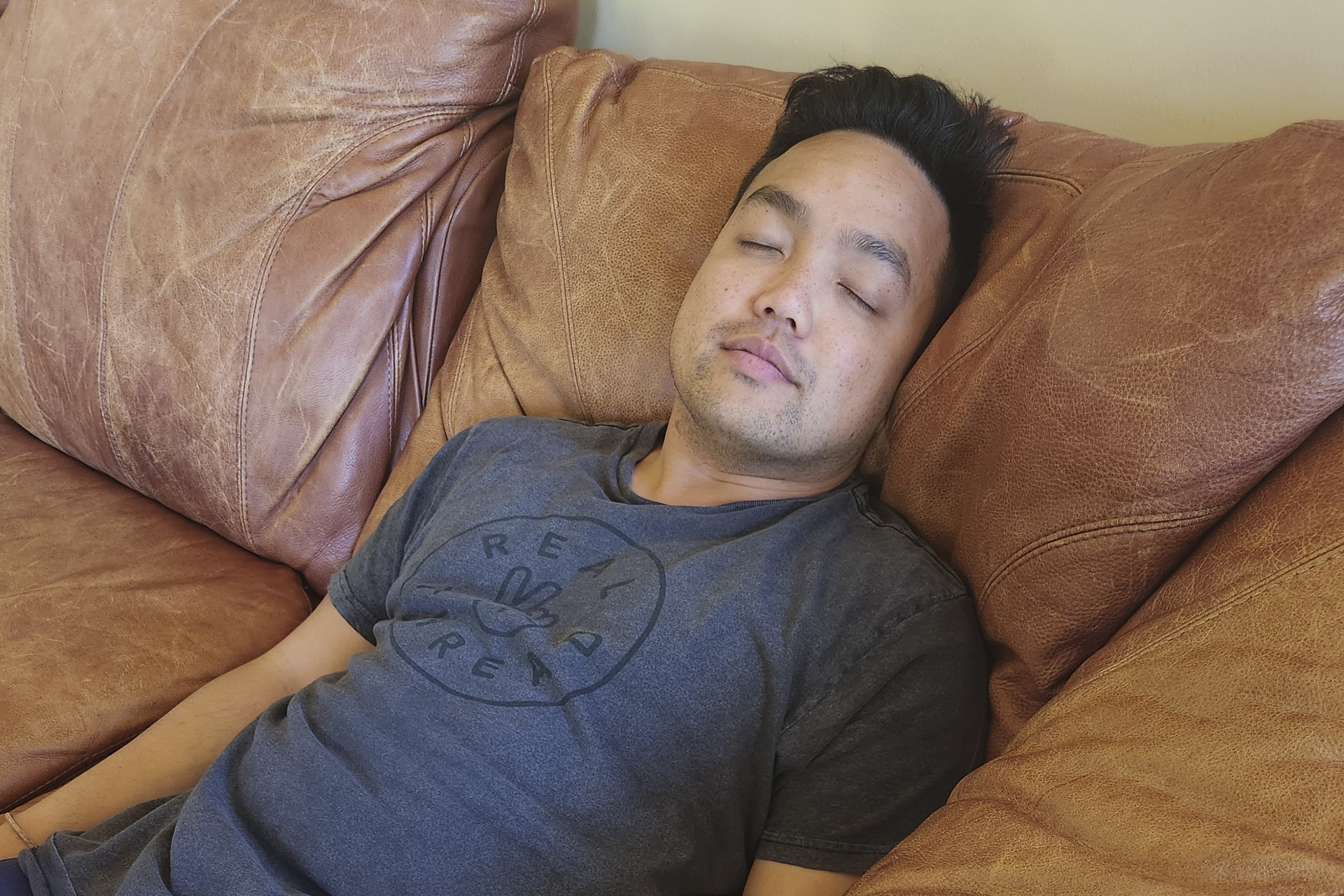 A young Asian man in a gray T-shirt rests on a brown leather couch with his eyes closed.