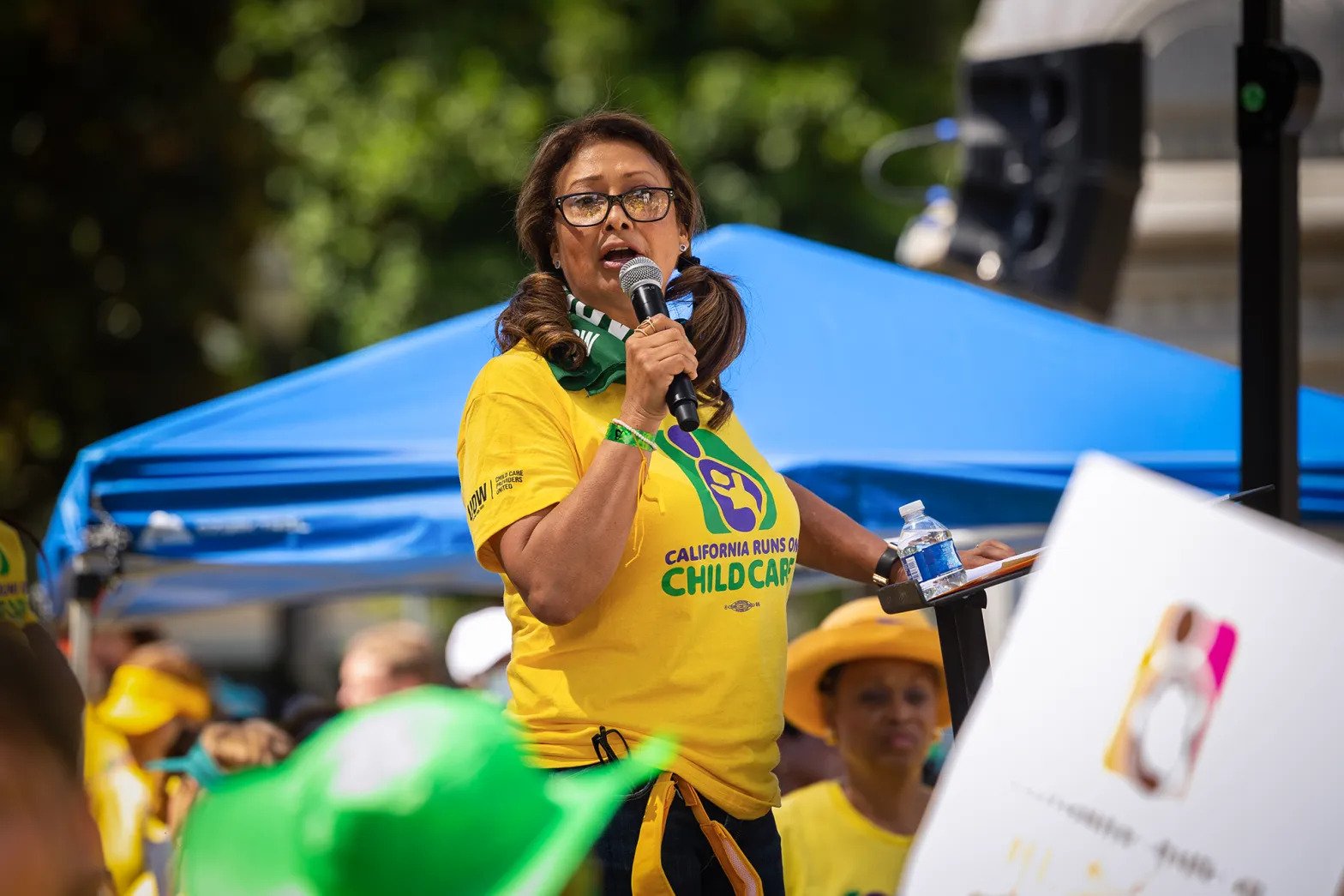 A Latina woman holding a microphone and wearing glasses stands in front of a crowd. She's wearing a yellow shirt.