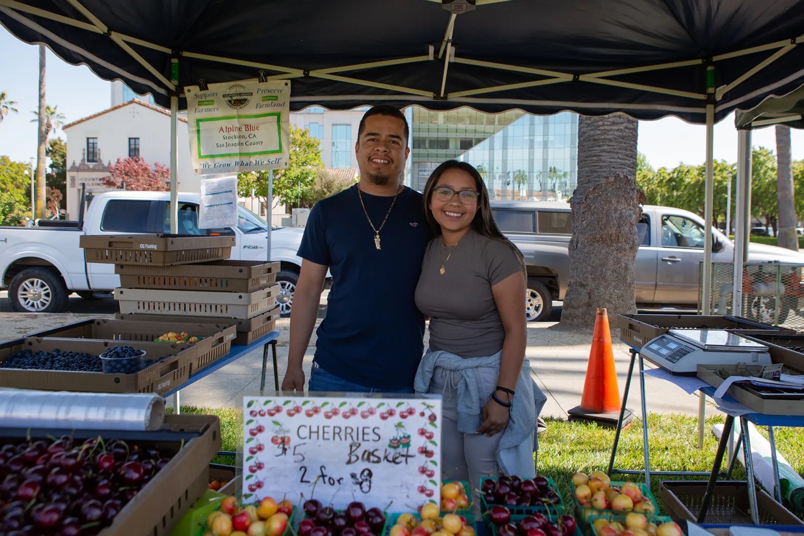 A Latino man and a Latino girl, both smiling and with arms around each other, stand under a tent and behind a table of fruit, with a sign advertising cherries