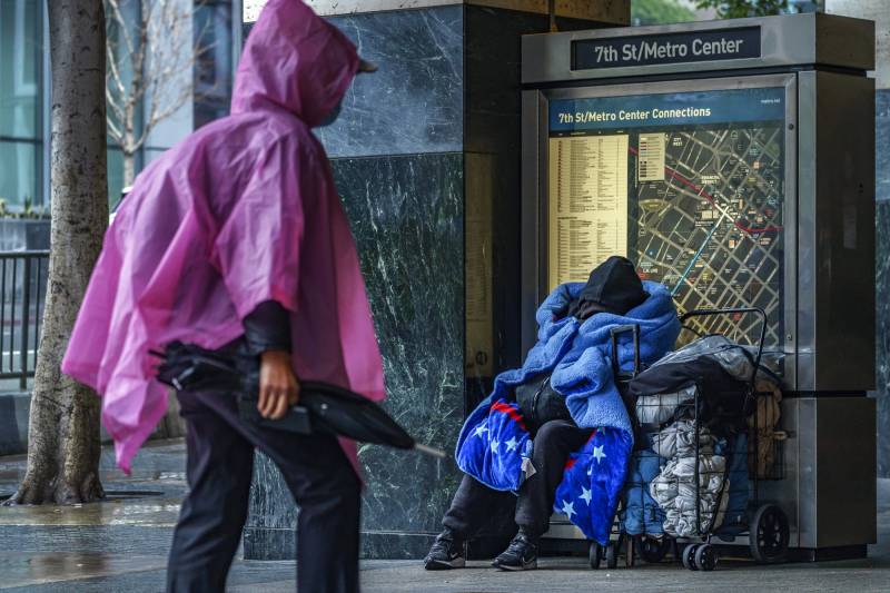 An unhoused person sits wrapped up in a blanket in front of a large metro map as another person wearing a pink poncho passes in front of them.