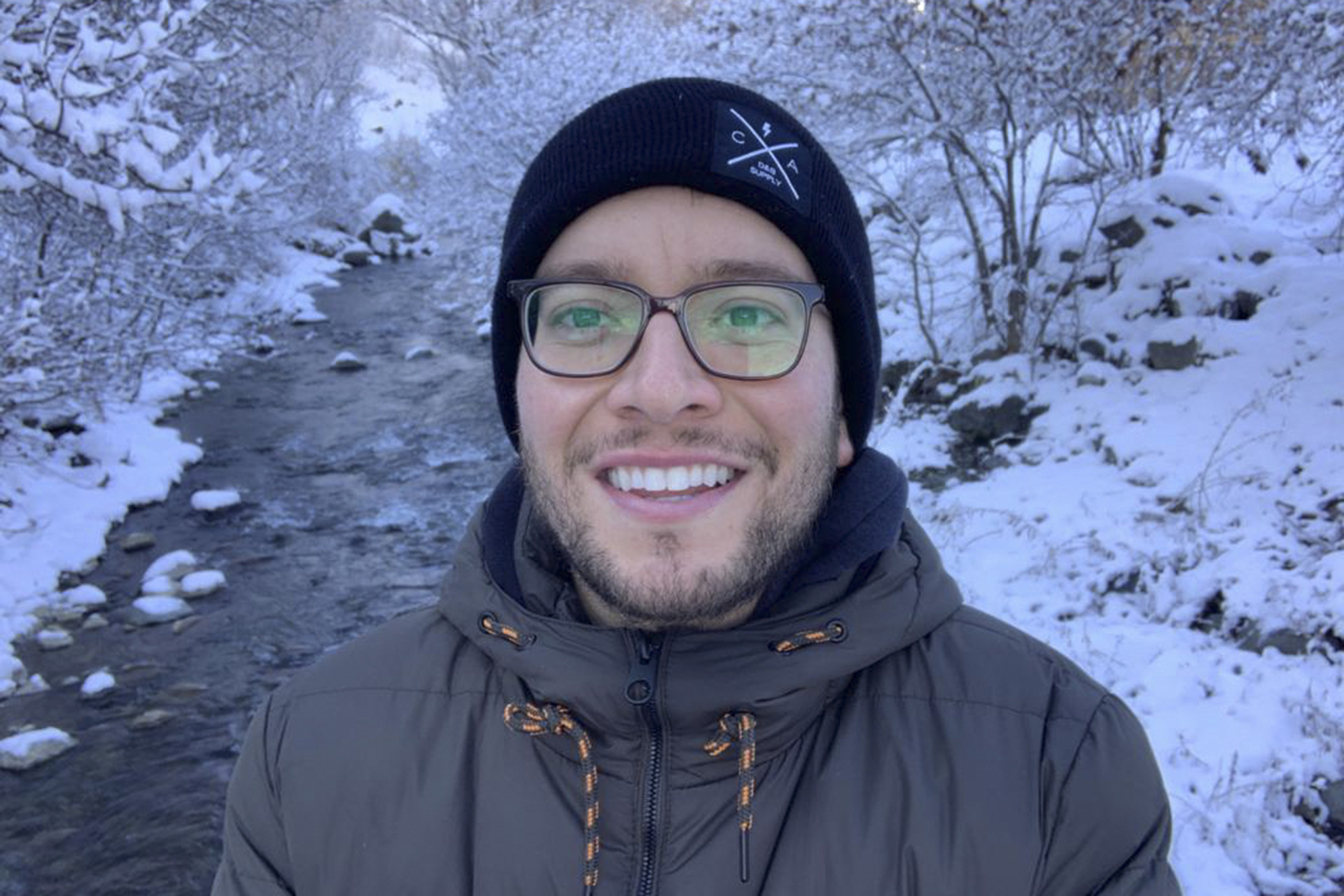 A person with short facial hair and glasses is photographed outdoors near a stream flowing through snowy terrain.