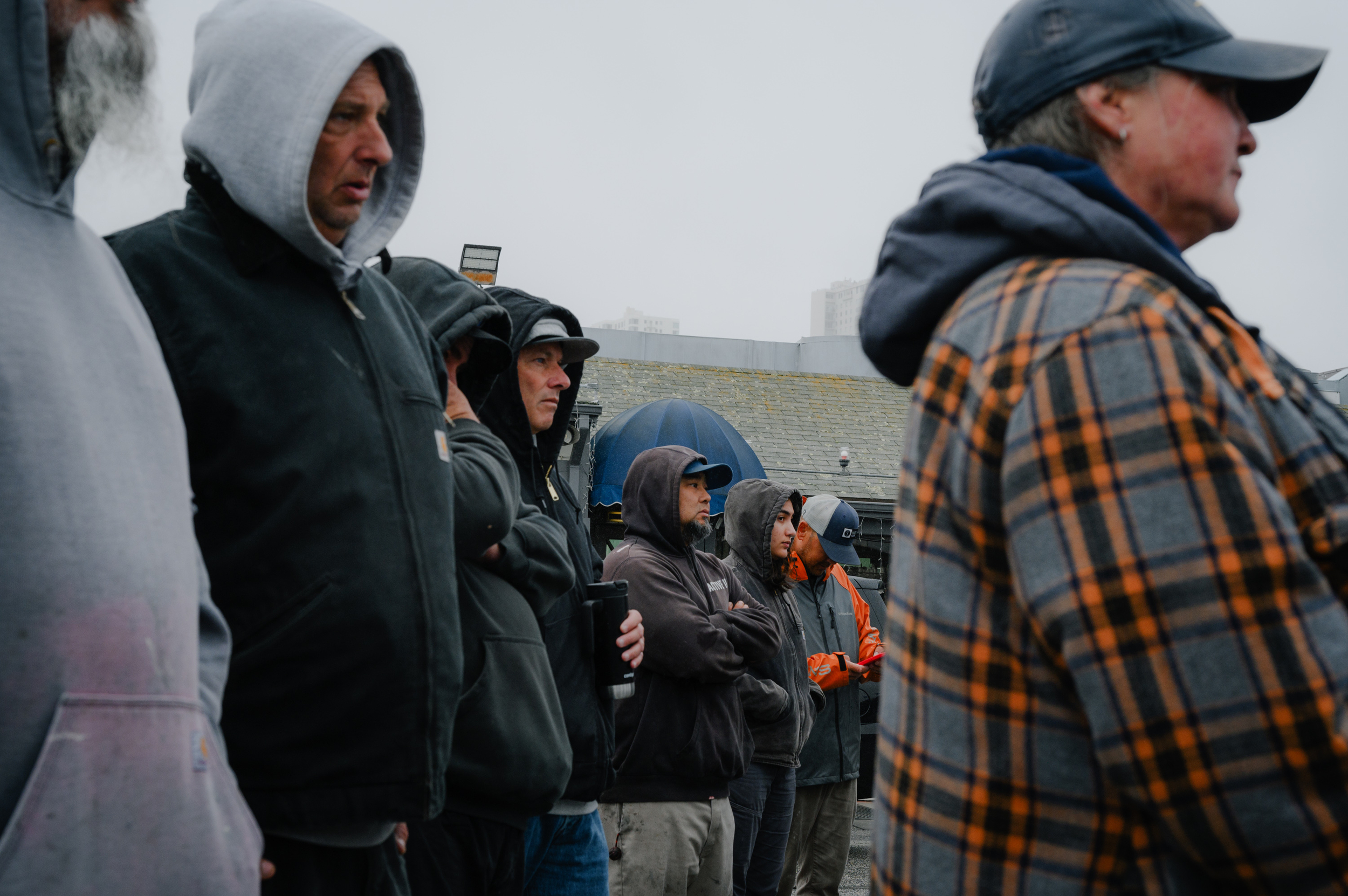 A group of people wearing jackets and sweatshirts with their hoods up and baseball caps all look in the same direction on a gray day.