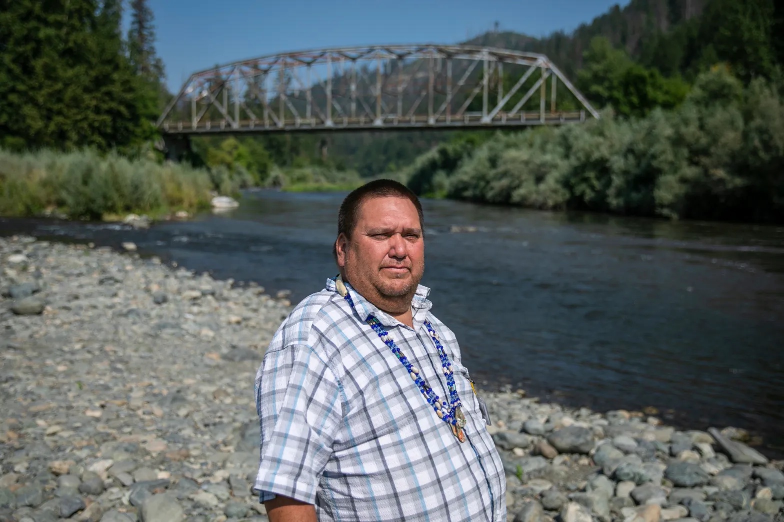 A larger Native man in a plaid button up shirt, with a long string of bright blue beads around his neck, stands in front of a river.